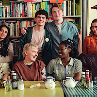A group of smiling young adults sits at a table with tea and condiments in a colorful cafe, with a bookshelf filled with books in the background, appearing relaxed and cheerful together.