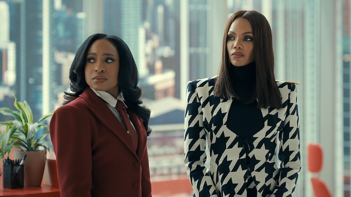 Two stylish people stand in a modern office with large windows showing a cityscape. One wears a red suit, the other a bold black and white jacket. They appear serious and attentive, surrounded by contemporary décor and plants.