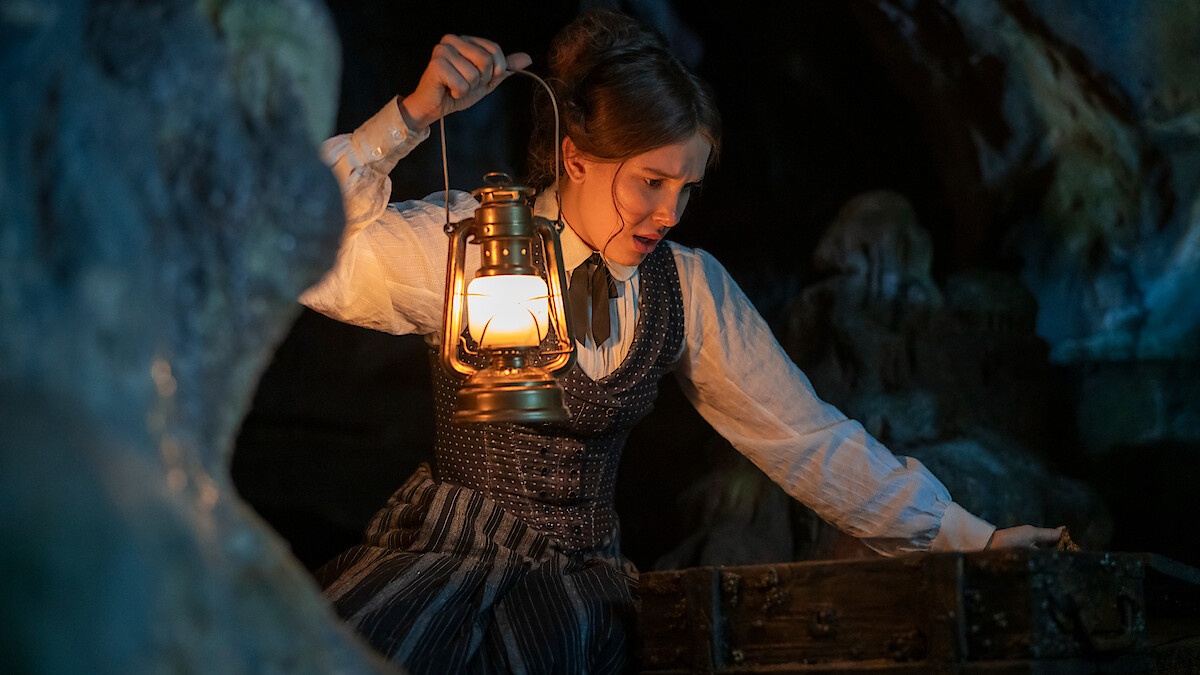 A woman in period clothing holds a lit lantern while exploring a dark, rocky cave, leaning over an old wooden chest, suggesting a search or discovery in a mysterious setting.