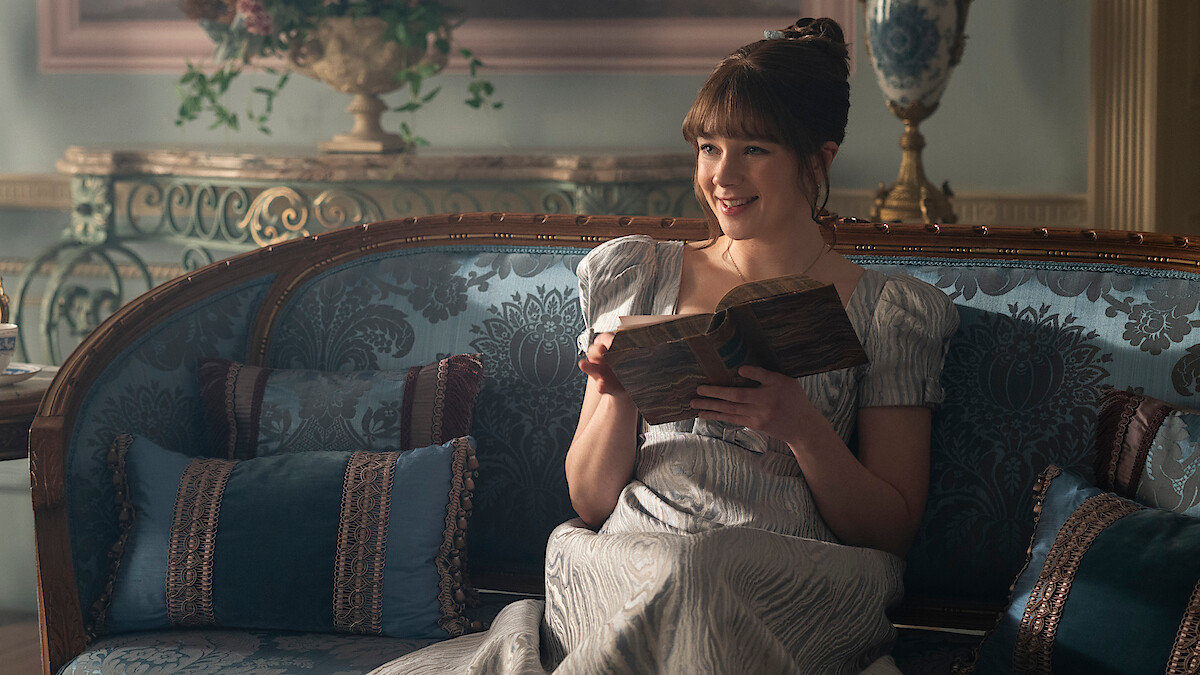 Woman in historical dress sitting on ornate blue couch in an elegant, vintage room, smiling while reading a book, surrounded by decorative cushions and classical furnishings.