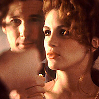 A close-up of a man and woman in a softly lit indoor setting, with the man in the background slightly out of focus and the woman in the foreground looking serious, holding a necklace near her neck.