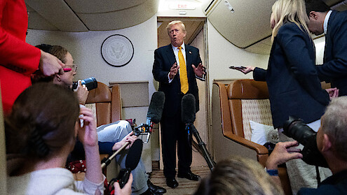 Man in suit speaks to reporters holding microphones and recording devices inside an airplane cabin, with leather seats and presidential seal visible in the background.