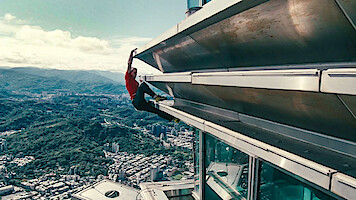 Alex Honnold in a red shirt climbing the exterior of a tall skyscraper above a city, with mountains and buildings visible in the distant background under a partly cloudy sky.