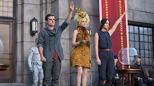 Three people stand in front of a stone building, two raising one hand; one wears a grey shirt, another a sleeveless outfit, and the third is in an elaborate orange dress at a microphone. Others are seated nearby, and a large banner hangs.