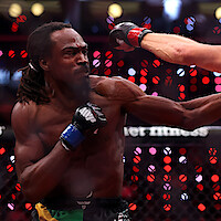 Two mixed martial artists fighting in a brightly lit cage, one landing a punch to the other's face during a match, with a blurred crowd and lights in the background.