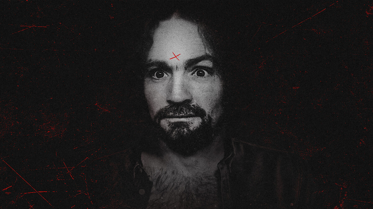 A man with curly hair and a beard stares intensely at the camera, set against a dark, gritty background. He has a red X marked on his forehead and visible chest hair, wearing an open shirt. The overall mood is eerie and dramatic.