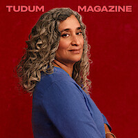 Geeta Gandbhir, director of The Perfect Neighbor, stands in front of a rich red backdrop with beautifully set curls in her graying hair, head turned toward the camera with a confident expression on her face.