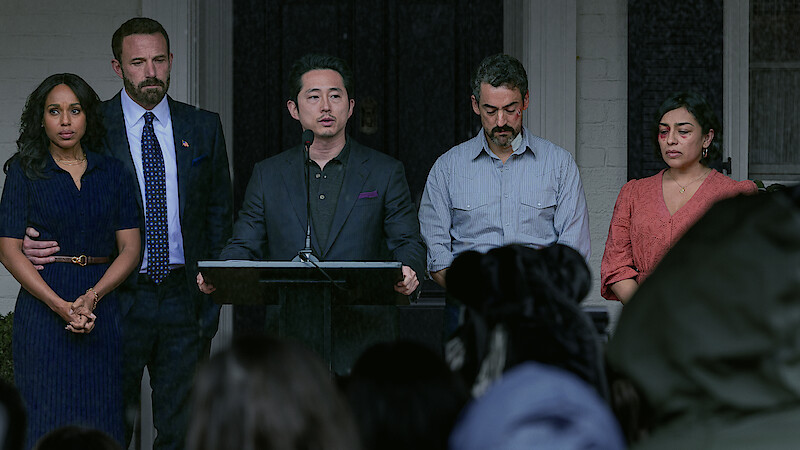 Five people stand together on a porch, one speaking at a podium with microphones, others looking serious; they face a crowd during what appears to be a press conference or public statement outdoors.