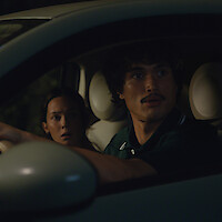 A man and woman sit in the front seats of a car at night, looking out the driver’s side window with serious and concerned expressions, illuminated by dim, moody lighting.
