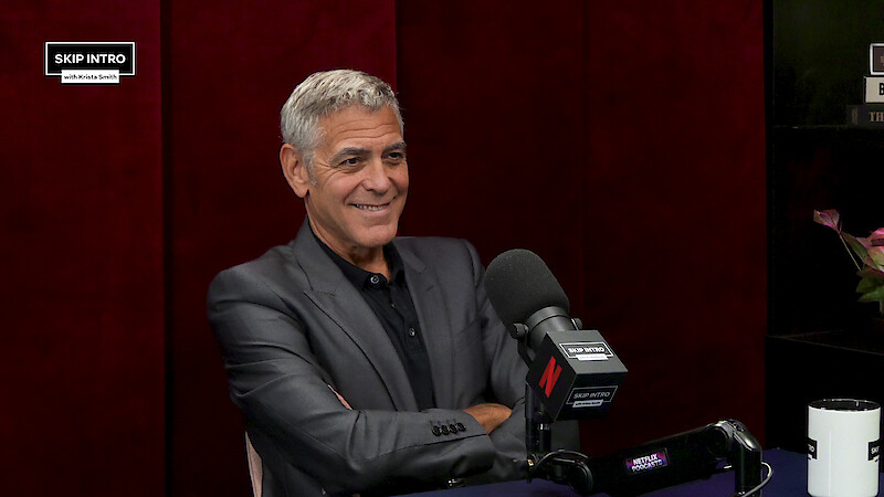 A man in a dark suit sits smiling at a podcast studio table, with a large microphone and "Skip Intro" branding, set against a red background. The mood is relaxed and professional.
