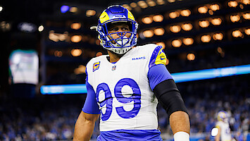 Football player in Los Angeles Rams uniform and helmet with number 99 stands on the field in a brightly lit stadium during a game, with a crowd visible in the background.