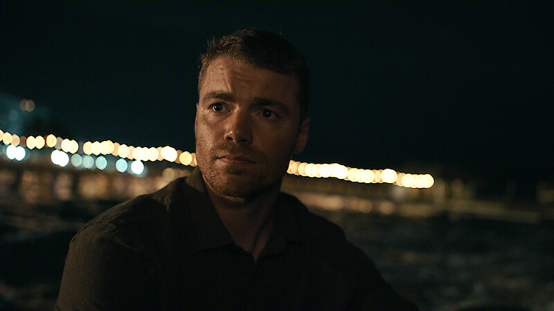 A man with short hair and a dark shirt sits outdoors at night near water, with a blurred background of lights from a pier or boardwalk reflecting on the surface.