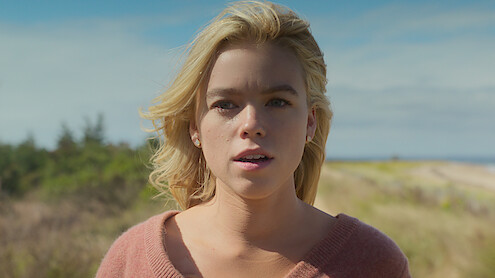 Milly Alcock wearing pink and standing on a beach in Sirens