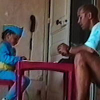 A child in a blue costume and adult sit at a small red table indoors, near a beige wall and a white door, engaged in an activity, with soft natural light in the room.