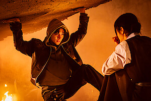 A person in a hoodie holds up a collapsing ceiling in a smoky, fire-lit room while another person crouches nearby, looking at them in concern.