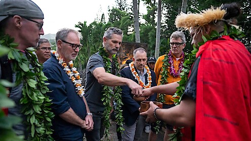 Group of people wearing leis and greenery participate in an outdoor traditional ceremony led by a person in red attire and a feathered headdress, surrounded by tropical trees.