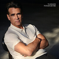 Colin Farrell leans back in a rolling chair in a white polo and black pants. 