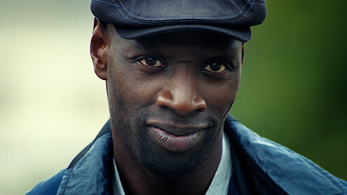 A man outdoors wearing a blue jacket, light shirt, and dark flat cap, looking directly at the camera with a subtle, confident smile and a blurred green background.
