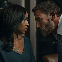 A serious man with a beard leans in closely toward a concerned woman in an indoor setting, both gazing intensely at each other, suggesting a tense or dramatic conversation.