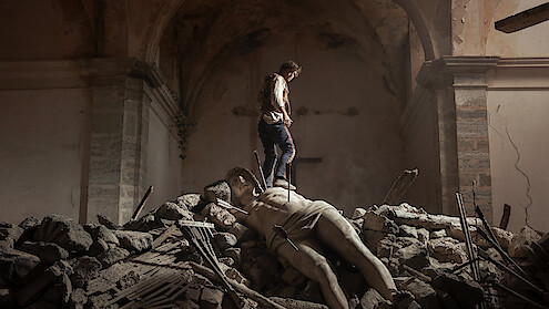 A person standing on rubble inside a damaged, old stone building, with a large fallen classical statue lying among the debris under an arched ceiling, evoking a sense of destruction and loss.