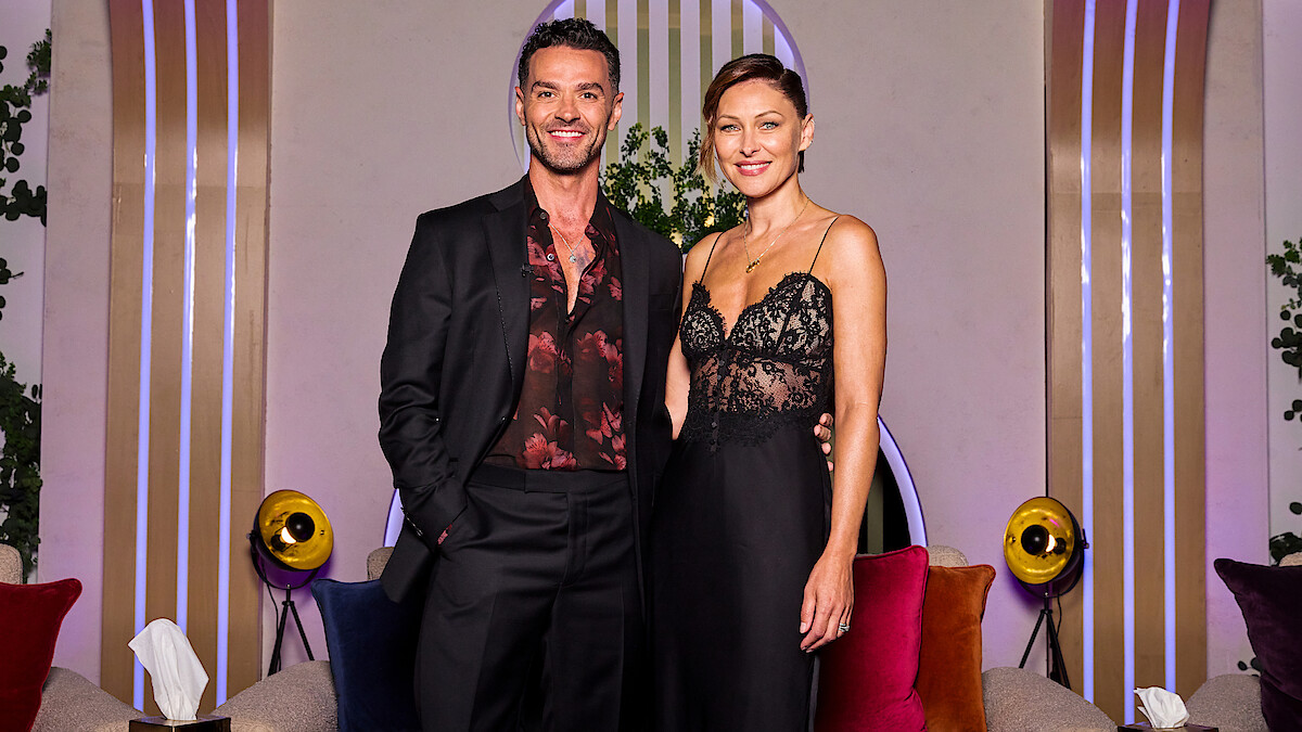 A man in a black suit and a woman in a black lace dress stand together smiling in a modern lounge with colorful cushions, round lights, plants, and decorative vertical lines in the background.