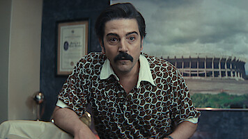 Man with dark hair and mustache in patterned shirt leans forward in an office with framed certificate on wall and large stadium photo in the background.