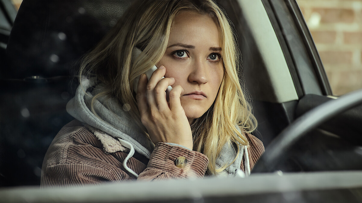 A young woman with blonde hair sits in a car, looking concerned while talking on a cellphone. She is wearing a hoodie and is positioned near the driver's seat, with a brick wall visible outside the window.