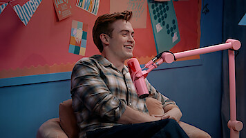 Kit Connor sitting and talking into a pink microphone in a colorful studio with artwork and notes on the wall behind them.
