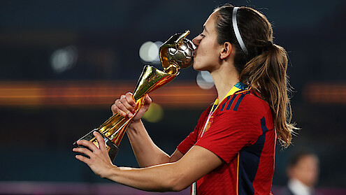 Soccer player in red jersey kisses gold trophy on stadium field, celebrating victory under stadium lights, wearing medal, hair tied back, background out of focus.