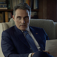 Man in a blue suit sitting in an armchair, holding papers, with bookshelves in the background, appearing serious in a formal office or library setting.