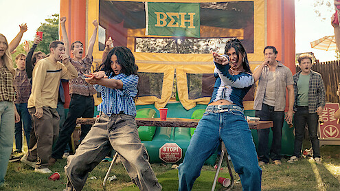 Two women dance energetically at an outdoor party with a crowd of cheering people, in front of a colorful bouncy house decorated with Greek letters, on a sunny day.