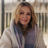 Woman with long blond hair wearing a cozy cream sweater and a thick purple-gray scarf, sitting outdoors with blurred fence and trees in the background on a soft, overcast day.