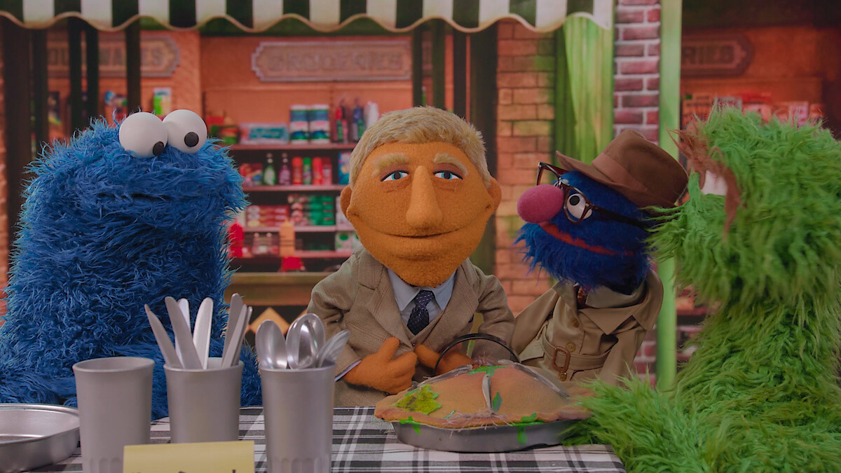 Four colorful puppets sitting at a checkered table in a cheerful, cozy café setting, with kitchen utensils and a pie.