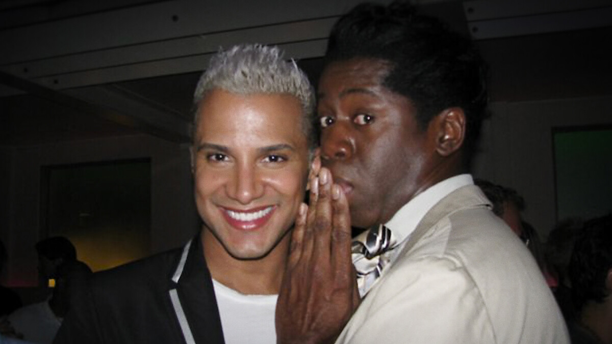 Two men in dressy attire pose indoors; one is smiling at the camera with platinum blond hair, while the other, wearing a bow tie, leans in to whisper with a playful expression. Dimly lit room with people and neutral colors in background.