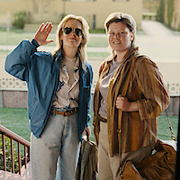 Two people standing outdoors on steps, dressed casually, one waving and wearing sunglasses, both carrying bags, with a residential or campus building and green lawn in the background on a sunny day.