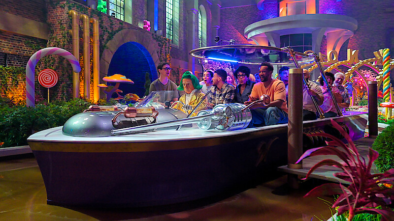 A group of people ride together in a boat through a colorful, whimsical indoor setting with candy decorations, glowing lights, lush plants, and fantasy architecture.