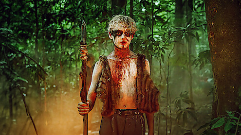 A person with painted face and fur vest stands holding a spear in a dense, misty, green forest, suggesting a primitive or survival setting.