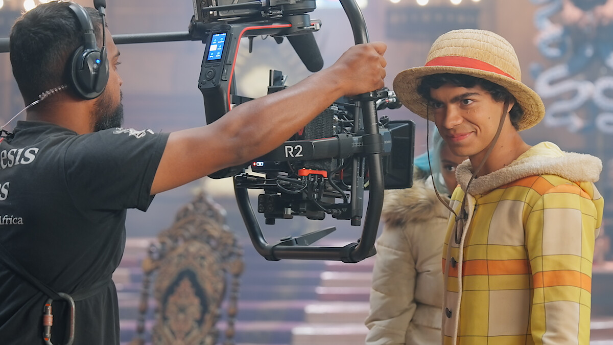 Iñaki Godoy as Monkey D. Luffy, in a yellow plaid costume and straw hat stands on a decorated film set, facing a professional camera rig held by a crew member, with cinematic lighting and ornate background elements visible.