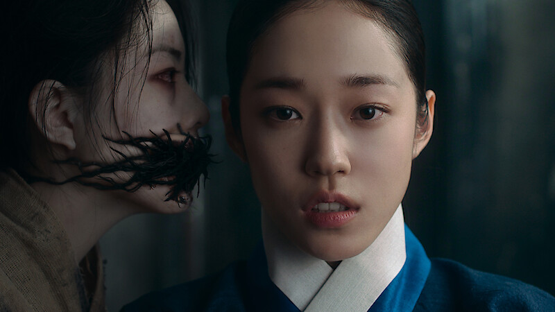 Two women indoors; one in traditional blue attire looks forward with a serious expression, while a ghostly figure with blackened mouth leans in closely from the side, creating a tense and eerie atmosphere.