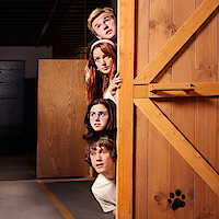 Four teenagers peeking curiously from behind a large wooden barn door with a paw print, in a warehouse-like setting with industrial lighting and wooden panels.