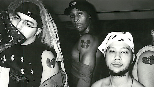 Three men pose in a black and white indoor photo, showing off Wu-Tang Clan tattoos and clothing featuring the group's logo. The environment is casual with urban streetwear and bandanas, emphasizing their affiliation and attitude.