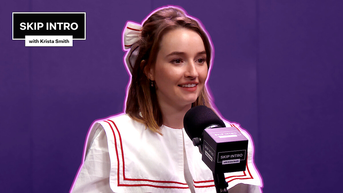 Kaitlyn Dever, smiling warmly, is seated in front of a microphone.