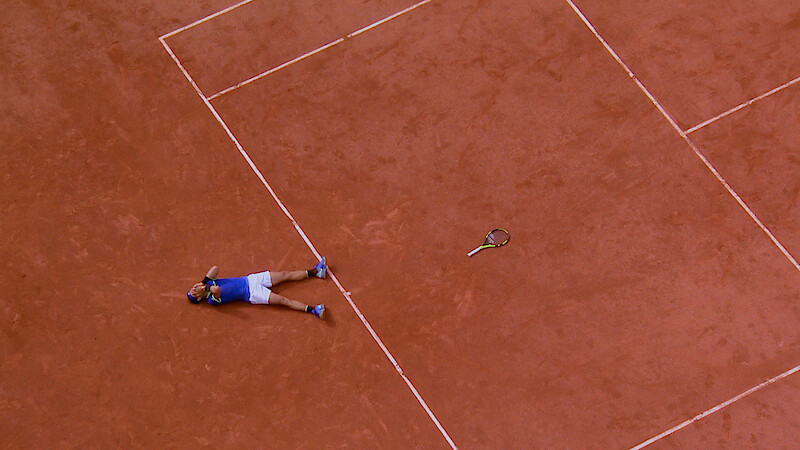 A tennis player lies on a clay court next to a tennis racquet, possibly celebrating or exhausted, viewed from above. The scene captures the lines and earthy texture of the tennis court.