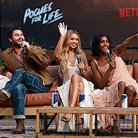 Three people sit on a couch holding microphones, smiling and waving, at a panel event with "POGUES FOR LIFE" and "NETFLIX" signs in the background. Cans and a coffee table are in front of them.
