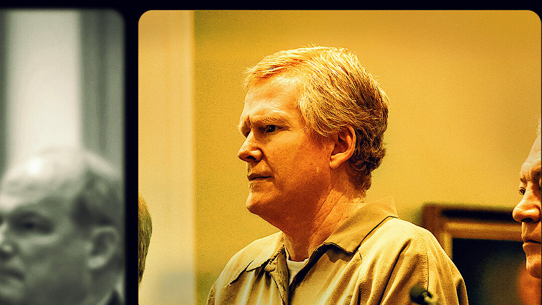 Middle-aged man with light hair in a tan prison uniform stands in a courtroom setting, with another man beside him and blurred figures in the background, all under warm indoor lighting.