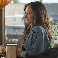 Minka Kelly as Quinn with long brown hair in a denim shirt sits at a table in a cozy diner, smiling, with large windows and a view of the street outside in the background.