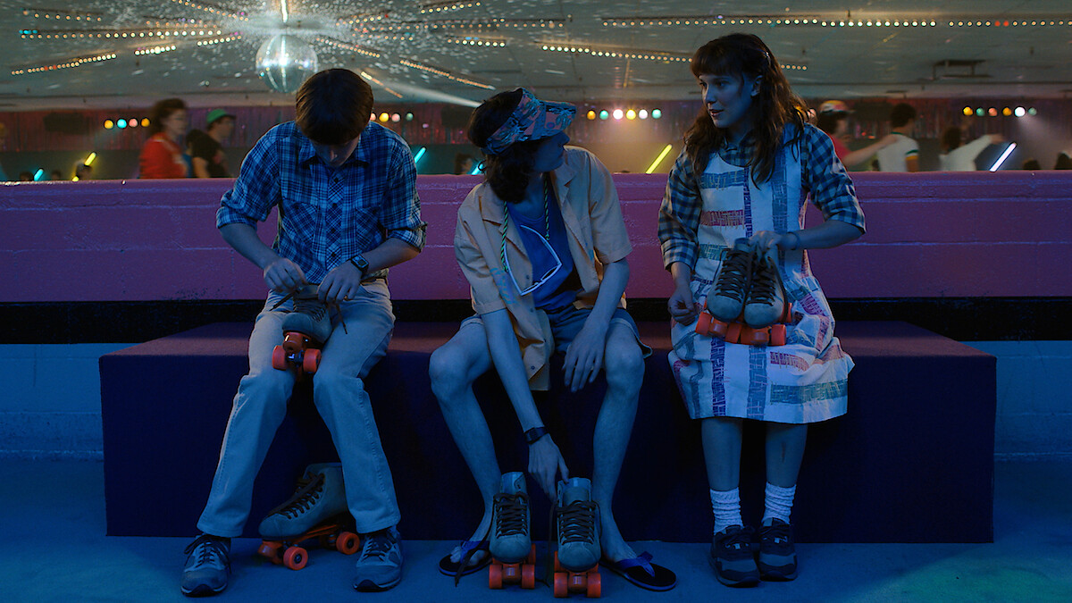 Noah Schnapp as Will Byers, Finn Wolfhard as Mike Wheeler and Millie Bobby Brown as Eleven tie shoes in a roller rink in 'Stranger Things' Season 4.