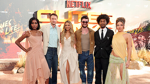 Six people pose together on a red carpet at an outdoor Netflix premiere event, dressed in fashionable attire, standing in front of a themed backdrop with desert elements and promotional text.