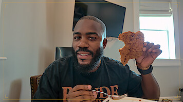 Man sitting indoors with a fork in one hand and holding a large piece of fried food in the other, smiling at the camera; modern room with a TV and window in the background.