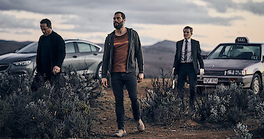Alex Dimitriades, Jamie Dornan, and Damon Herriman stand in a desert landscape with two cars in the background in 'The Tourist'